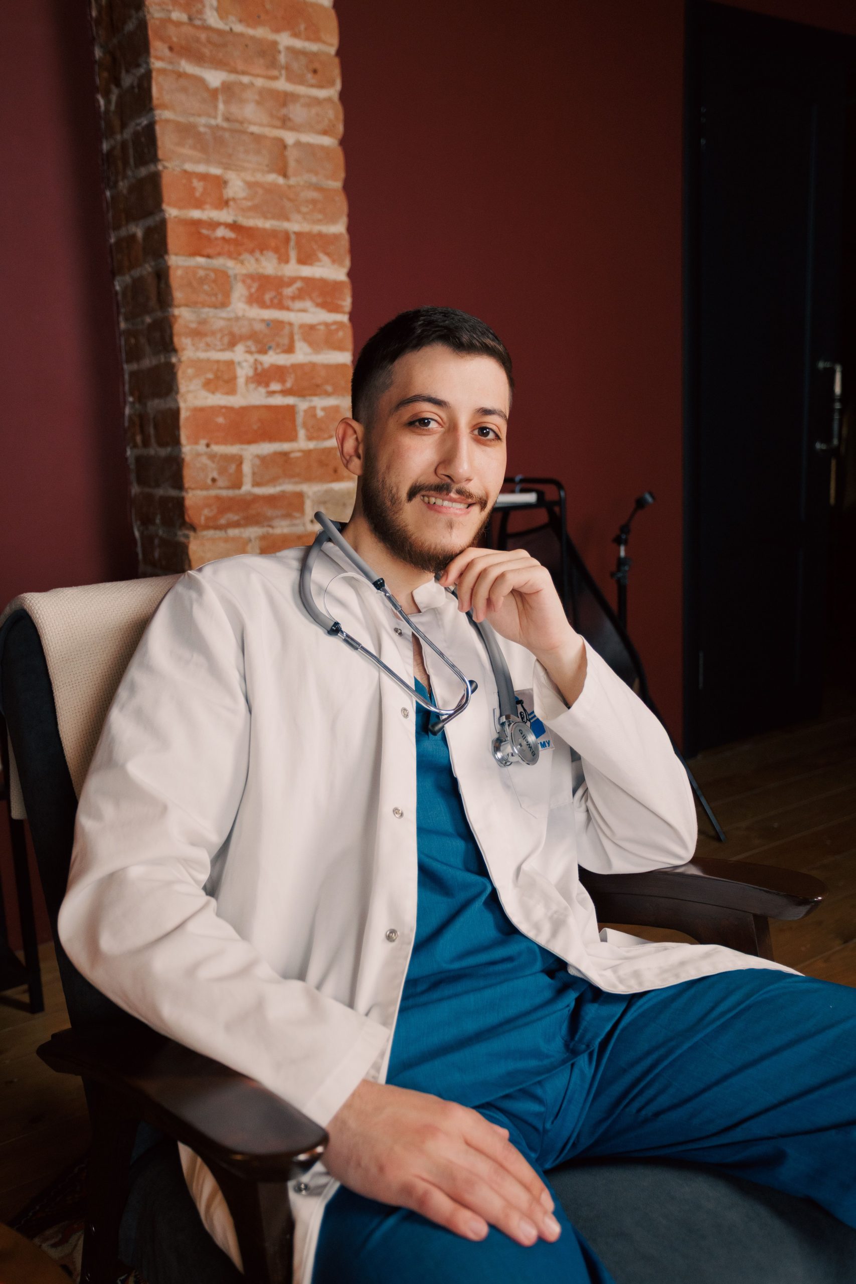 Young physician seated in a medical office wearing blue scrubs and a white lab coat with a stethoscope, representing professionals looking to Apply for Medical Practice Loan, secure doctor practice purchase loans, obtain dermatology practice loans, compare healthcare loan companies, and explore medical technology leasing solutions for practice growth and expansion.